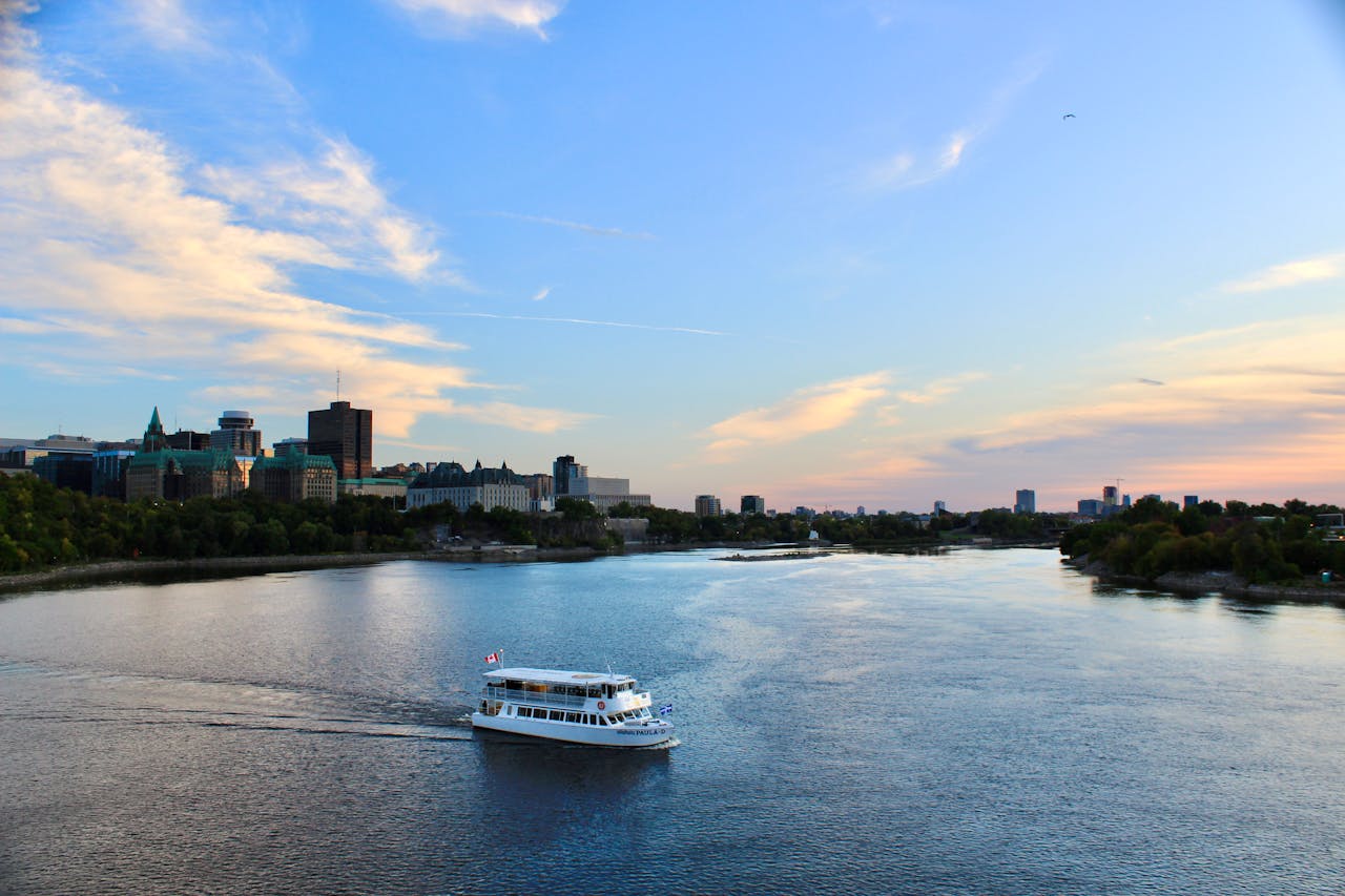 A touring boat cruises on a river with a city skyline backdrop under a bright sky.